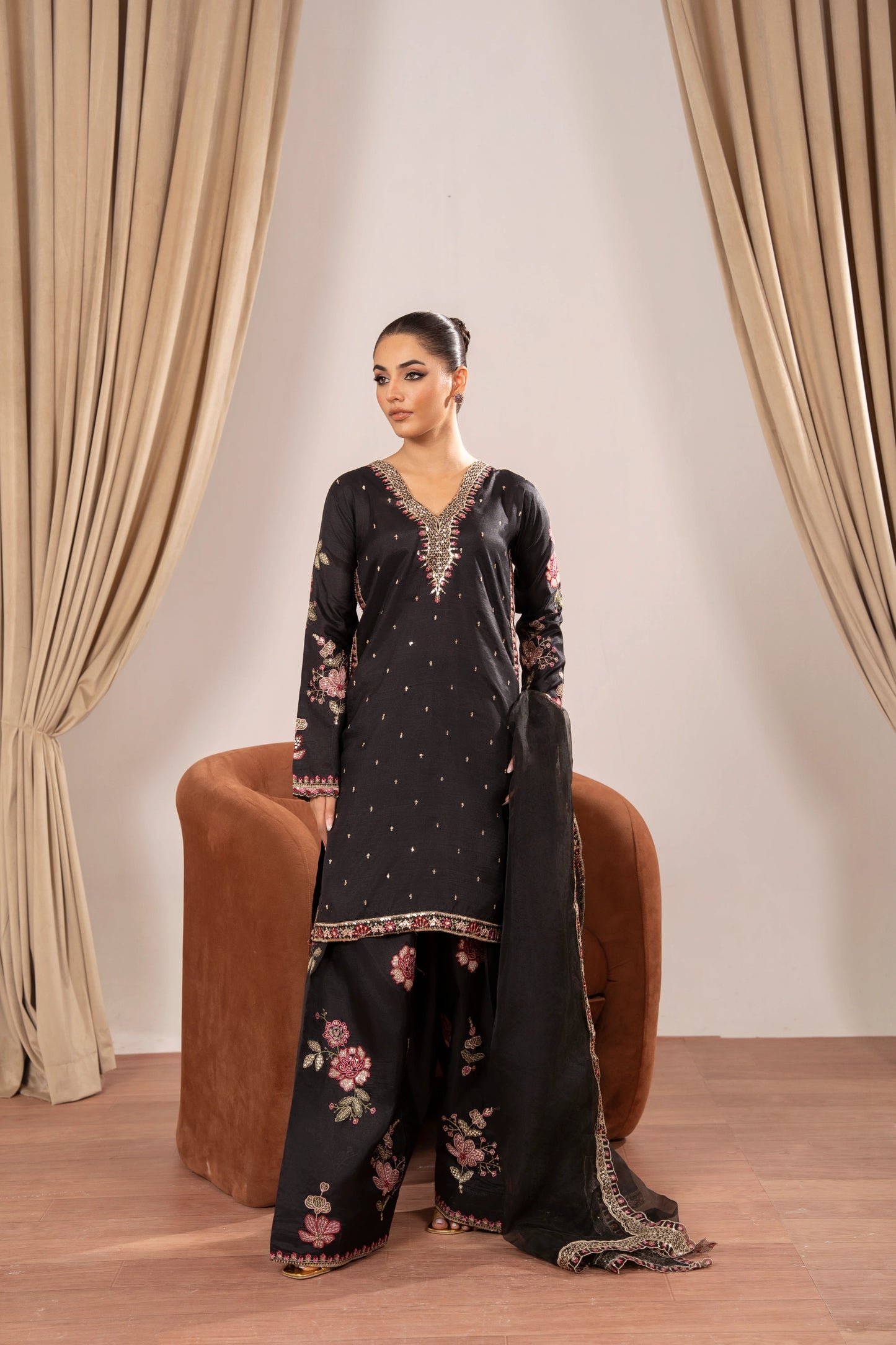 Woman in a black embroidered outfit sitting on a brown chair with beige curtains in the background