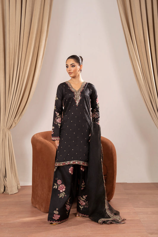 Woman in a black embroidered outfit sitting on a brown chair with beige curtains in the background
