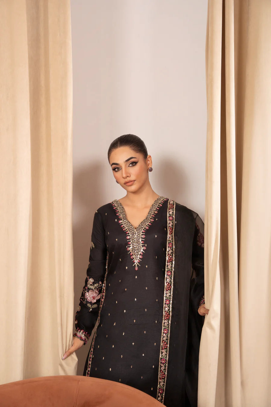 Woman wearing a black embroidered traditional outfit standing between beige curtains.