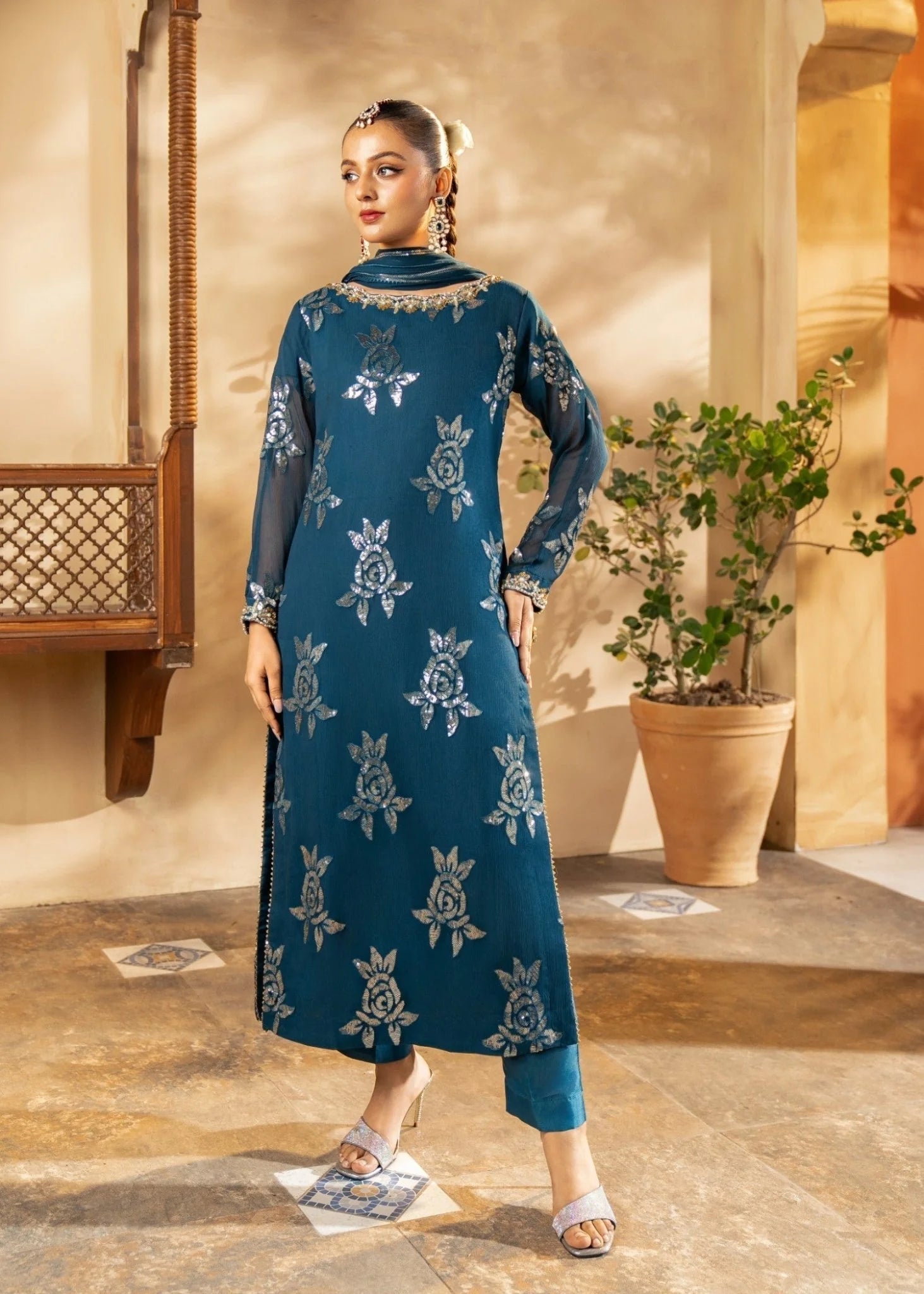 Woman in a blue embroidered dress standing in an indoor setting with plants and architectural elements.