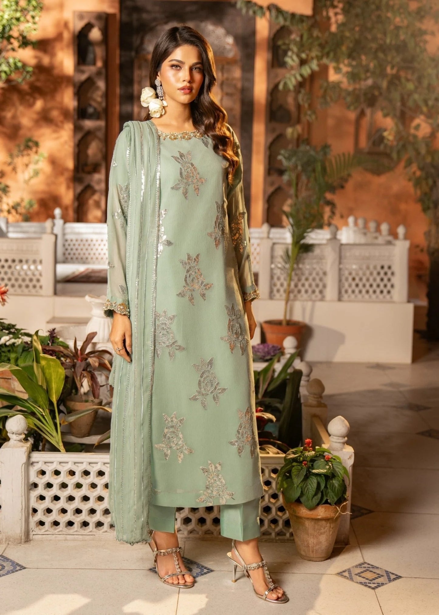 Woman in a light green embroidered outfit standing in an outdoor setting with plants and furniture.