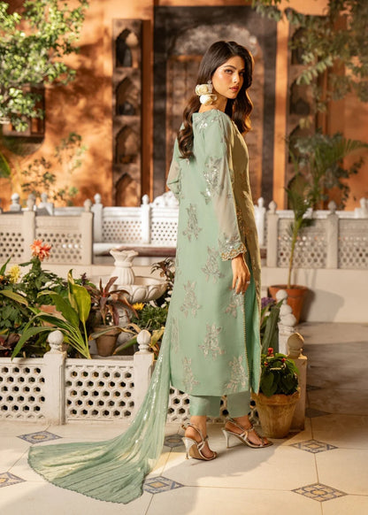 Woman in a light green traditional outfit standing outdoors with plants and a building in the background