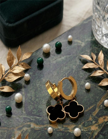 Gold hoop earrings with black clover charms on a dark surface with pearls and leaves.
