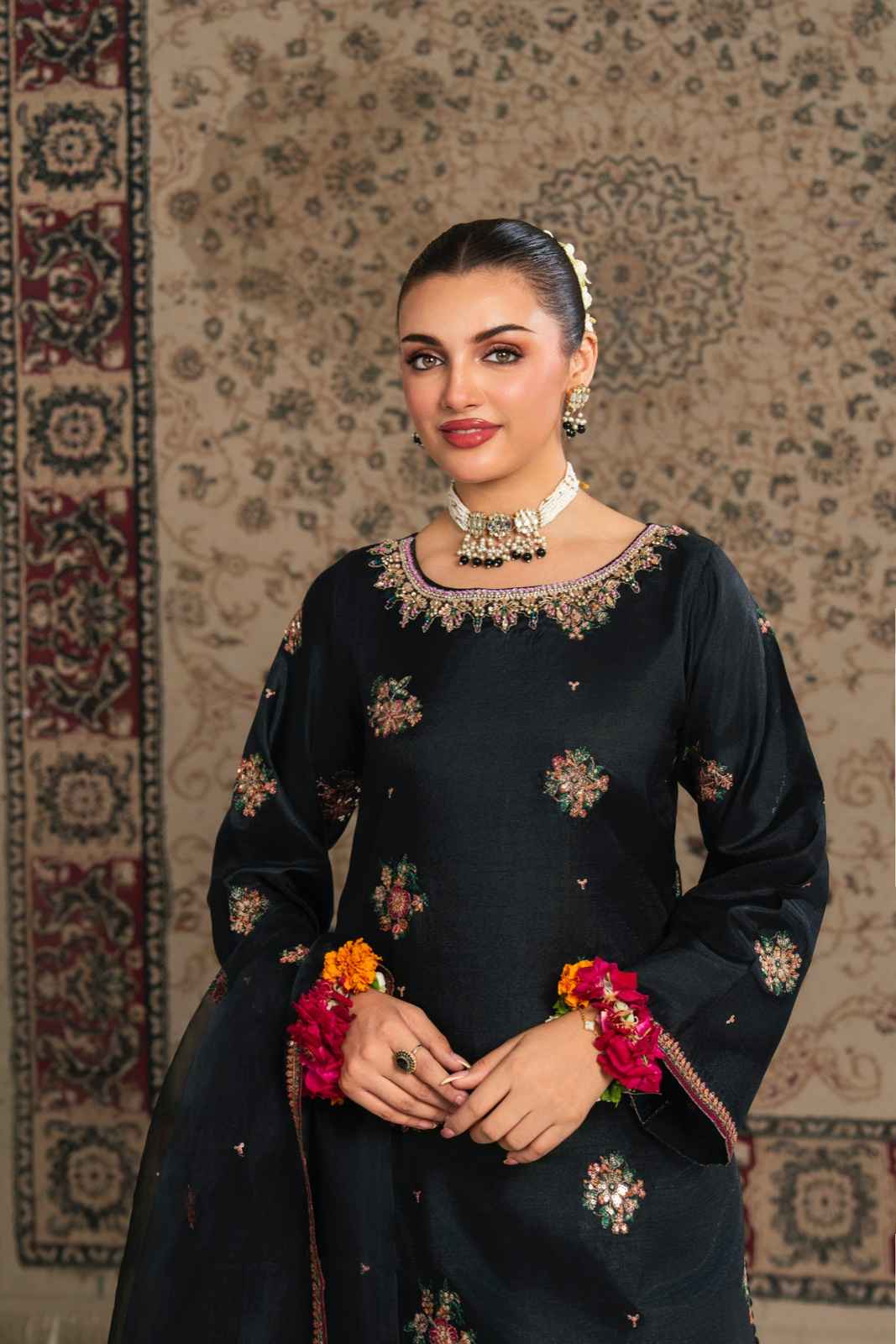 Woman in a black traditional outfit with floral patterns and jewelry, standing against a patterned wall.