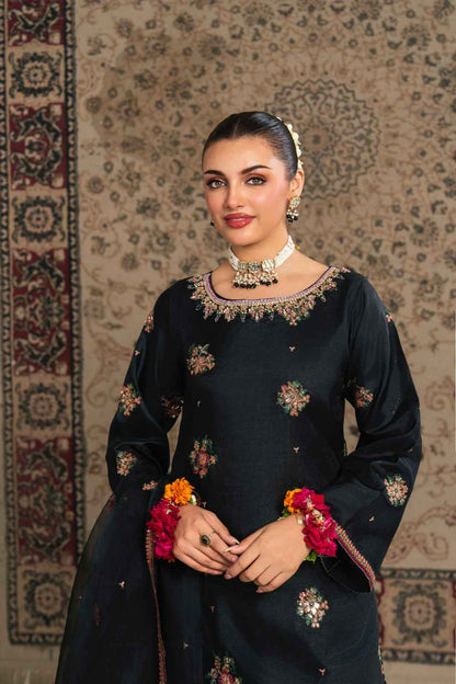 Woman in a black traditional outfit with floral patterns and jewelry, standing against a patterned wall.