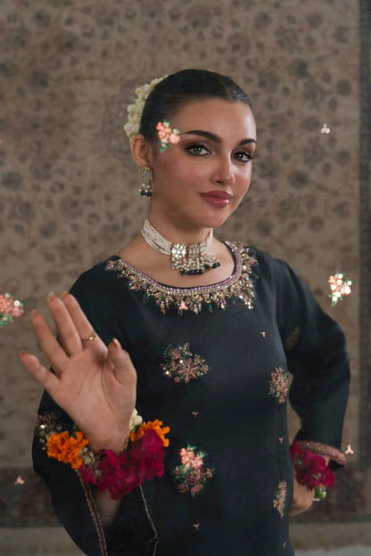 Woman in traditional attire with floral decorations, posing against a patterned wall.