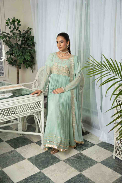 Woman in a light green traditional outfit standing in a room with white curtains and a checkered floor.