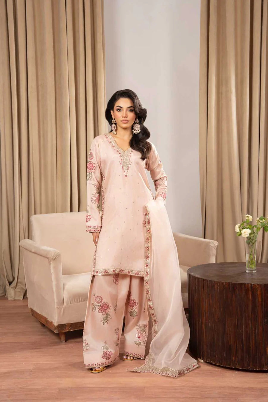 Woman in a pink traditional outfit standing in a room with beige curtains and a wooden table.