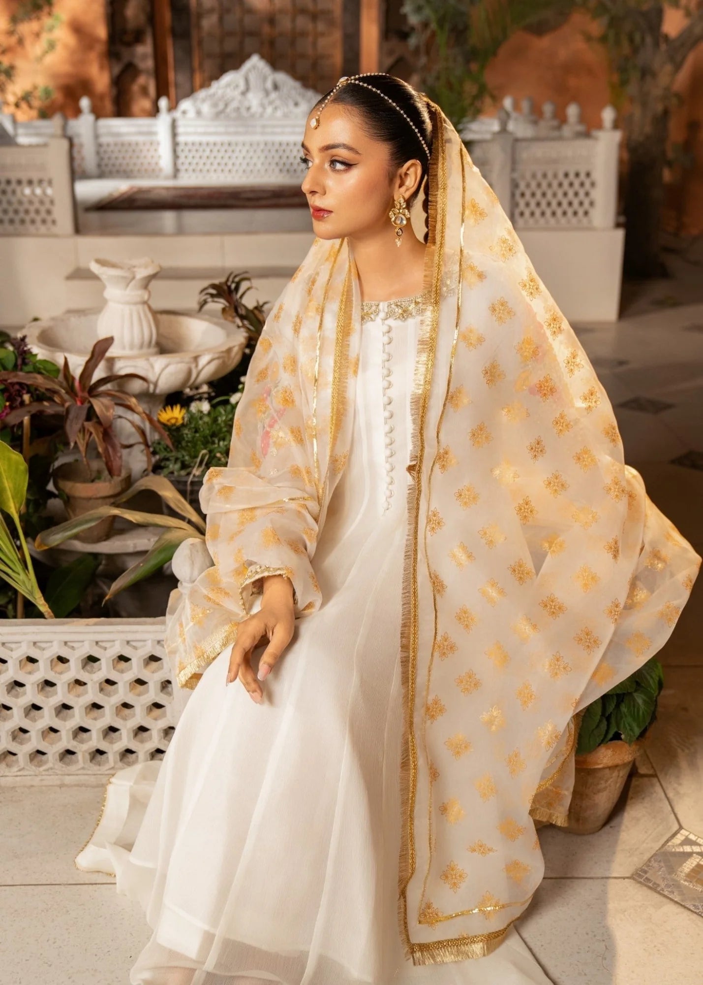 Woman in a white and gold traditional outfit sitting outdoors with plants and a fountain in the background.